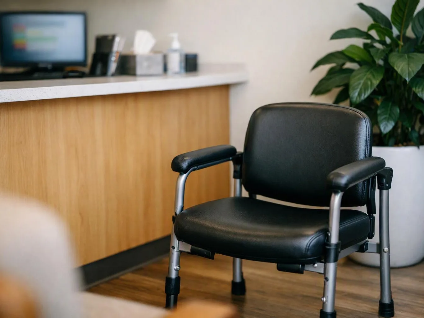 A black padded chair sits in a waiting room beside a counter, with a potted plant in the corner